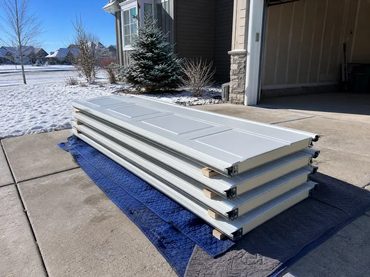 New insulated garage door sections stacked on moving blankets in a snowy Marquette driveway on install day